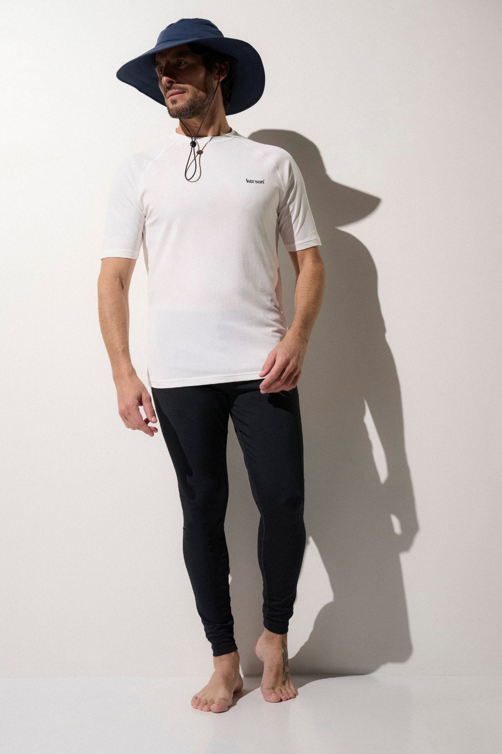 Homme en chapeau anti-UV bleu, t-shirt blanc UPF 50+ et leggings noirs, idéal pour la protection solaire lors d'activités en extérieur.