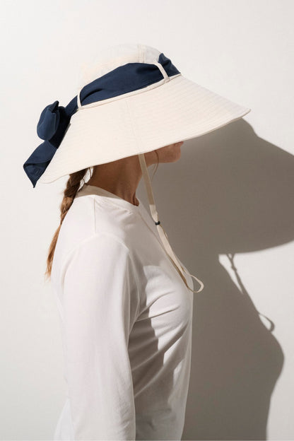 Chapeau anti-UV femme blanc, avec large bord pour une très haute protection solaire, porté avec un ruban bleu marine.